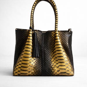 May include: Black and gold tote bag with a textured snakeskin pattern. The bag features a structured design with a rounded handle and a decorative tassel. The gold sections have black stripes.