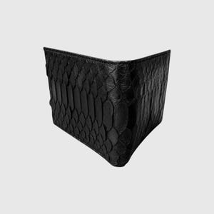 Könnte beinhalten: Eine schwarze Geldbörse im Bifold-Design mit einer strukturierten Schlangenhaut-Optik auf der Außenseite. Die Geldbörse ist leicht geöffnet dargestellt und zeigt die ausgeprägten schuppenartigen Details sowie die dunkle, matte Oberfläche.