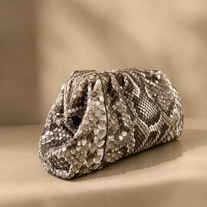 May include: A structured clutch bag featuring a textured snakeskin pattern in shades of brown, beige, and cream. The bag has a gathered, pouch-like silhouette with a soft, rounded shape and a concealed frame closure, showcasing a detailed scale texture.