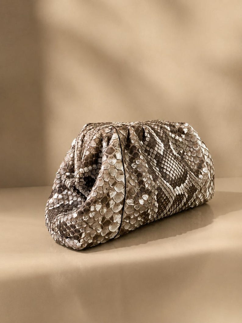May include: A structured clutch bag featuring a textured snakeskin pattern in shades of brown, beige, and cream. The bag has a gathered, pouch-like silhouette with a soft, rounded shape and a concealed frame closure, showcasing a detailed scale texture.