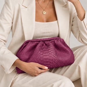 Pode incluir: Uma bolsa clutch magenta profunda com um padr&atilde;o de pele de cobra texturizado e um design franzido de estrutura macia. O acess&oacute;rio tipo bolsa tem uma silhueta arredondada e um fecho de dobradi&ccedil;a, exibindo uma cor rica, saturada e textura.