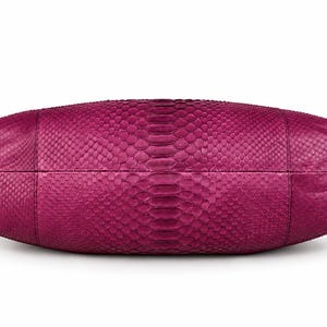 Pode incluir: Uma bolsa de m&atilde;o magenta com um padr&atilde;o de pele de cobra texturizado em toda a sua superf&iacute;cie. A bolsa tem uma forma oval horizontal alongada com costuras vis&iacute;veis dividindo os pain&eacute;is, exibindo uma textura de escamas consistente por toda parte.