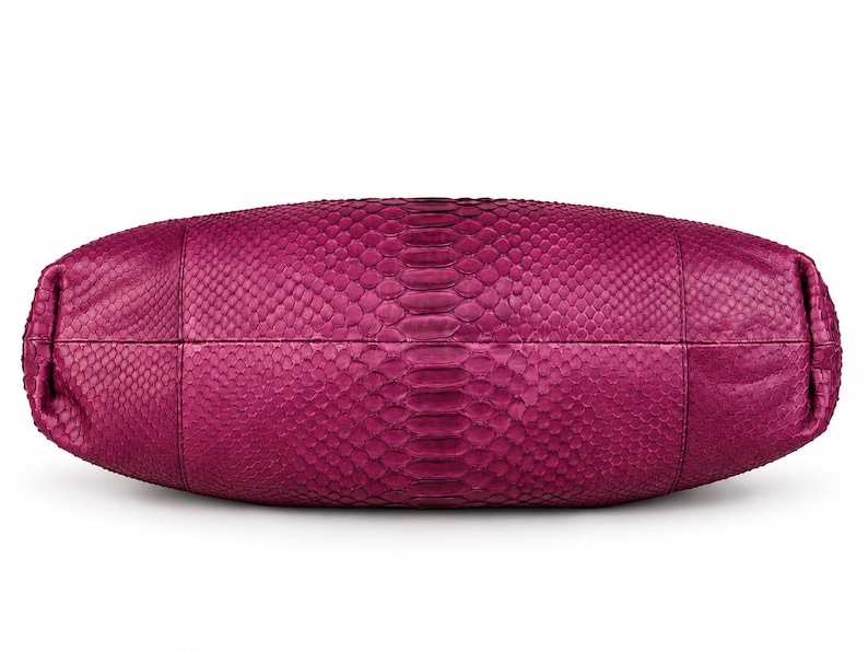 Pode incluir: Uma bolsa de m&atilde;o magenta com um padr&atilde;o de pele de cobra texturizado em toda a sua superf&iacute;cie. A bolsa tem uma forma oval horizontal alongada com costuras vis&iacute;veis dividindo os pain&eacute;is, exibindo uma textura de escamas consistente por toda parte.