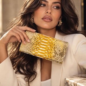 May include: A rectangular clutch bag featuring a vibrant yellow and metallic gold snakeskin-textured pattern. The accessory has a structured, elongated shape with a flap closure and a distinct scale-like surface finish throughout the exterior.