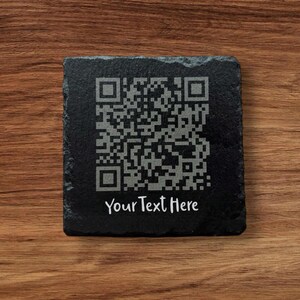 May include: A square black slate coaster featuring a QR code and the text "Your Text Here" in white. The coaster is displayed on a wooden surface.
