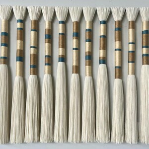 May include: A wall hanging made of white yarn with brown and teal stripes. The yarn is tied together at the top and hangs down in a fringe.