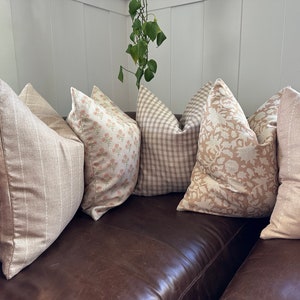 May include: Five decorative throw pillows in various patterns and colors, including beige, brown, and pink, arranged on a brown leather couch.