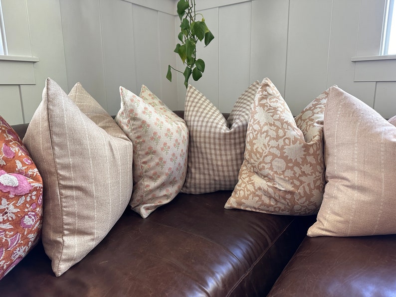 May include: Five decorative throw pillows in various patterns and colors, including beige, brown, and pink, arranged on a brown leather couch.
