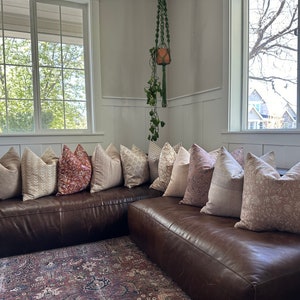 May include: A brown leather sectional sofa with a variety of patterned throw pillows in shades of beige, pink, and red. The sofa is positioned in front of a window with a view of trees and houses.
