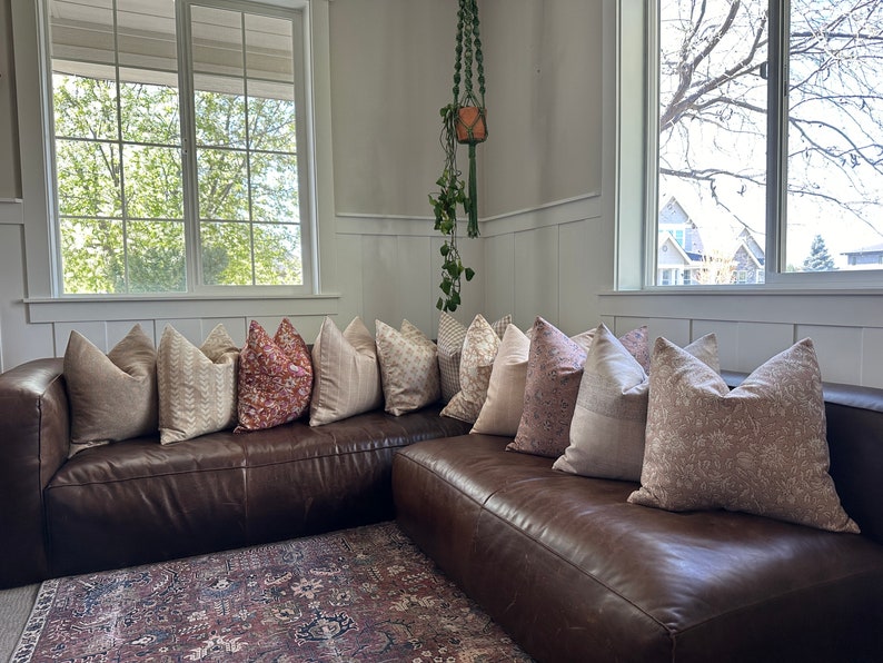 May include: A brown leather sectional sofa with a variety of patterned throw pillows in shades of beige, pink, and red. The sofa is positioned in front of a window with a view of trees and houses.