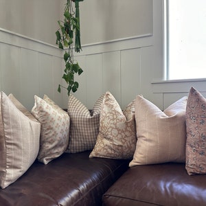 May include: A collection of throw pillows in various shades of beige and pink with different patterns, including floral, geometric, and striped designs. The pillows are arranged on a brown leather couch.