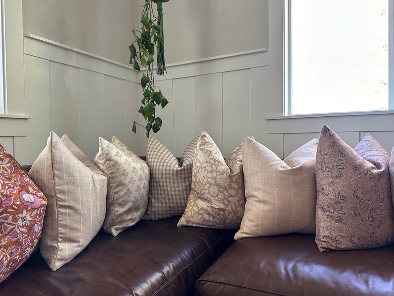 May include: A collection of throw pillows in various shades of beige and pink with different patterns, including floral, geometric, and striped designs. The pillows are arranged on a brown leather couch.