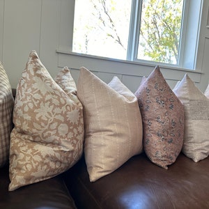 May include: Five decorative throw pillows with different patterns in shades of brown and beige. The pillows are arranged on a brown leather couch.