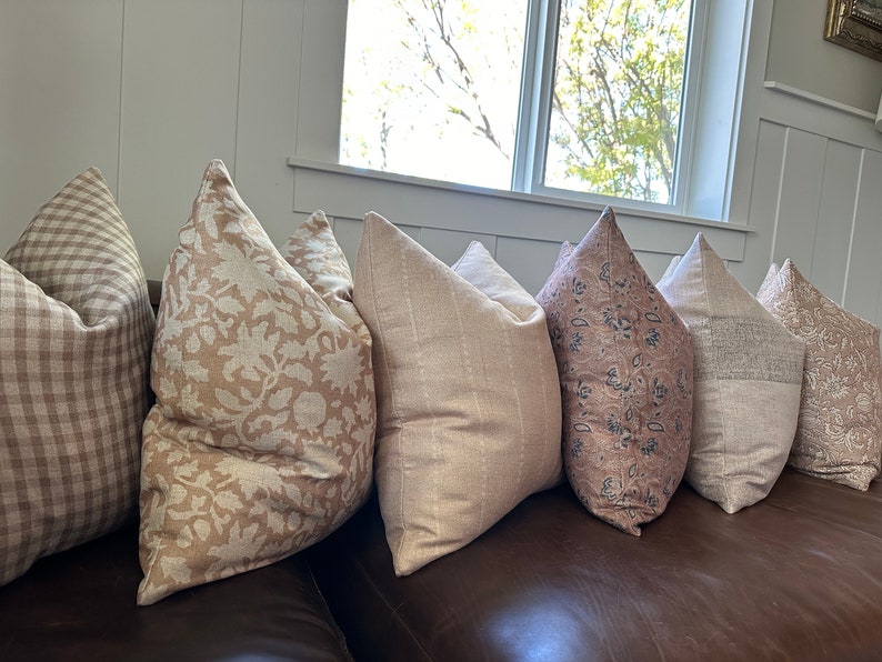 May include: Five decorative throw pillows with different patterns in shades of brown and beige. The pillows are arranged on a brown leather couch.