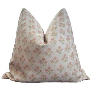 Puede incluir: Un cojín cuadrado con una base de tela beige claro que presenta un patrón repetitivo de pequeños grupos de flores de color rosa suave y tallos de color verde apagado. El cojín tiene un aspecto estructurado y relleno con un pliegue central.