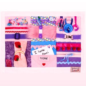 May include: A pink sensory board with multiple textures and interactive features. Includes zippers, buttons, a pop-it toy, a buckle, and a small envelope with a card reading "I love nani" and the name "Yvonne".
