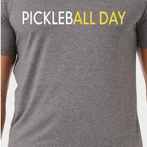Può includere: Una maglietta grigia con il testo "PICKLEBALL DAY" in lettere bianche e gialle.