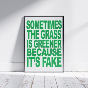 May include: A framed poster with the text "SOMETIMES THE GRASS IS GREENER BECAUSE IT'S FAKE" in bright green letters. The poster is in a black frame and leans against a white wall on a white wooden floor.