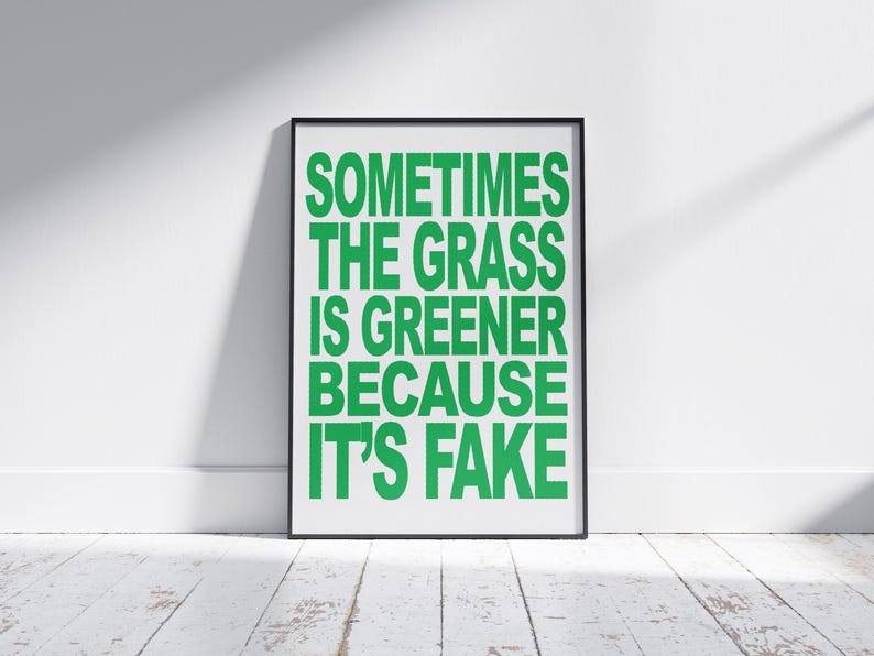 May include: A framed poster with the text "SOMETIMES THE GRASS IS GREENER BECAUSE IT'S FAKE" in bright green letters. The poster is in a black frame and leans against a white wall on a white wooden floor.