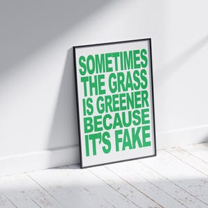 May include: A framed print with the text "SOMETIMES THE GRASS IS GREENER BECAUSE IT'S FAKE" in bright green letters. The black frame is leaning against a white wall and a white wooden floor.