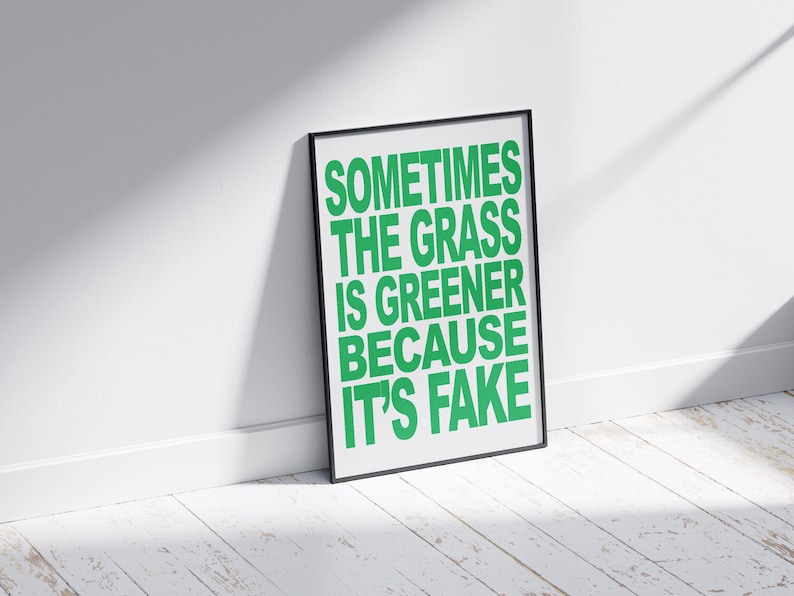 May include: A framed print with the text "SOMETIMES THE GRASS IS GREENER BECAUSE IT'S FAKE" in bright green letters. The black frame is leaning against a white wall and a white wooden floor.