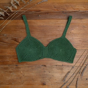 Op de afbeelding: Een groen gecrochete crop top met een cirkelvormig patroon. De top heeft dunne bandjes en ligt plat op een houten oppervlak.