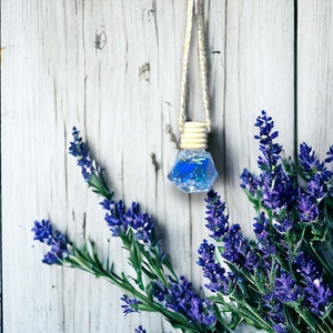 May include: A blue glass bottle with a diamond shape hangs from a string. The bottle is filled with a blue liquid and glitter. The bottle is surrounded by lavender flowers.
