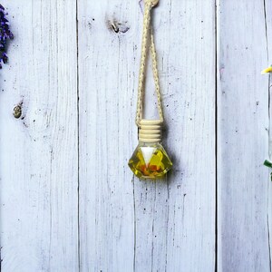 May include: A yellow glass bottle with a string hanging from it. The bottle is filled with a yellow liquid and has a wooden cap. The bottle is hanging on a white wooden background. There are purple flowers on the left and white flowers on the right.