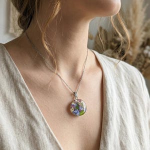 Puede incluir: Un collar plateado con un colgante redondo lleno de flores secas coloridas. El colgante, de aproximadamente 2,5 cm de diámetro, cuelga de una cadena delicada. Las flores incluyen flores moradas, azules y blancas.