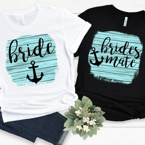 Może przedstawiać: Dwie koszulki, jedna biała i jedna czarna, z motywami marynistycznymi. Biała koszulka ma napis "Bride" z czarną kotwicą, a czarna koszulka ma napis "Brides Mate" z czarną kotwicą. Oba wzory znajdują się na jasnoniebieskim tle z fakturą drewna.