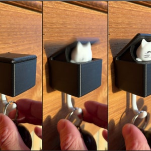 Może przedstawiać: Czarny uchwyt na klucze z otwieraną pokrywą, zamontowany na haczyku ściennym. Wewnątrz pudełka znajduje się biała figurka kota. Pokrywa otwiera się, odsłaniając kota. Uchwyt na klucze jest przymocowany do drewnianych drzwi.