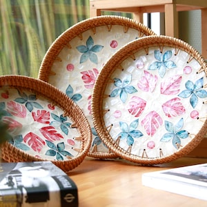 Peut inclure: Trois plateaux ronds en osier avec des motifs floraux blancs, roses et bleus. Les plateaux sont faits de matériaux naturels et ont un aspect rustique.