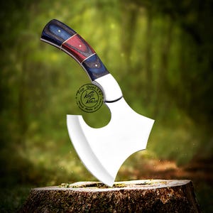 May include: A hand-made axe with a blue, red, and black wooden handle. The axe head is made of stainless steel and has a hole in the center. The axe is resting on a tree stump. The text "Top Quality Knife Factory Hand Made Knives" is printed on the handle.