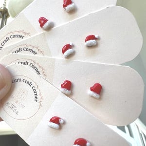 May include: A set of six red and white Santa hat earrings. The earrings are displayed on a white card with the text "Curti Craft Corner".