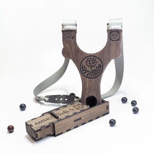 Może przedstawiać: Drewniana proca z szarym paskiem i małym drewnianym pudełkiem z etykietą "Ammo" z tekstem "Trifles by KLIC" na boku. Proca ma wygrawerowany wzór drzewa i pola na przodzie.