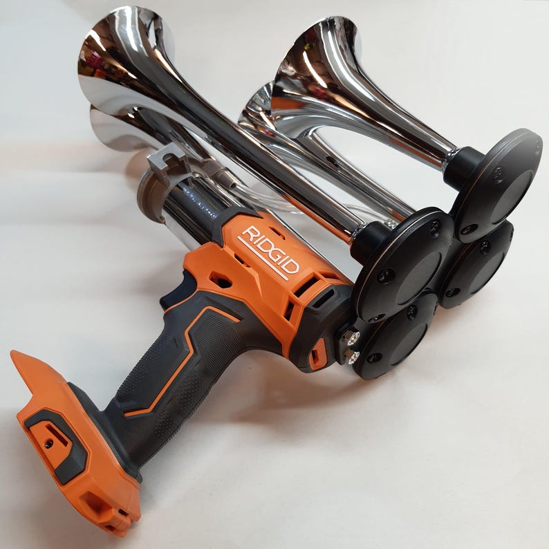 Op de afbeelding: Een zwart en oranje draadloos gereedschap met een RIDGID-logo. Het gereedschap is bevestigd aan vier chromen luchttoeters.