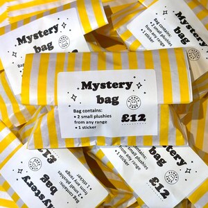 May include: A yellow and white striped paper bag with a white label that reads Mystery bag, Bag contains: 2 small plushies from any range, 1 sticker, and a small circular logo with a ghost icon. The label also features small star symbols.