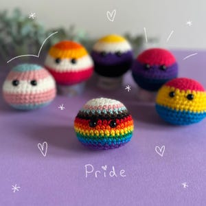 May include: A selection of crocheted spheres, each representing a different LGBTQ+ pride flag. The spheres are in various colours, including rainbow, pink, blue, yellow, and purple. The word "Pride" is written in white script on a purple background.