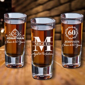 Puede incluir: Tres vasos de chupito de vidrio transparente con grabados personalizados. El primer vaso dice "50 Jonathan Cheers to 50 Years". El segundo vaso dice "M Miguel Aged to Perfection". El tercer vaso dice "60 Johnson Cheers to 60 Years".