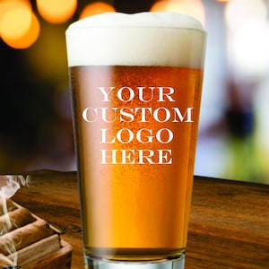 May include: A pint glass filled with golden beer and a thick white head. The glass has the text "YOUR CUSTOM LOGO HERE" in white. The background is blurred with warm, out-of-focus lights, and the glass sits on a wooden surface.
