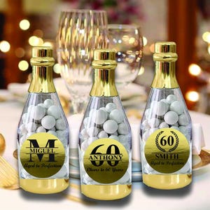 May include: Three gold champagne bottle favors filled with white candy. Each bottle has a personalized gold sticker with a name, age, and a celebratory message. The stickers say "Miguel Aged to Perfection", "Anthony Cheers to 60 Years", and "Smith Aged to Perfection".