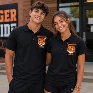 May include: Two people wearing black polo shirts with the Pleasantville Tigers logo. The logo features a tiger's head in orange and white. The shirts have a collared neckline and short sleeves. The background includes a brick wall with the words "TIGER PRIDE".