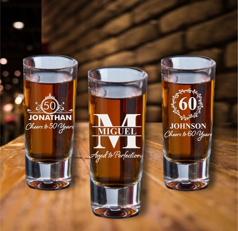 Puede incluir: Tres vasos de chupito de vidrio transparente con grabados personalizados. El primer vaso dice "50 Jonathan Cheers to 50 Years". El segundo vaso dice "M Miguel Aged to Perfection". El tercer vaso dice "60 Johnson Cheers to 60 Years".