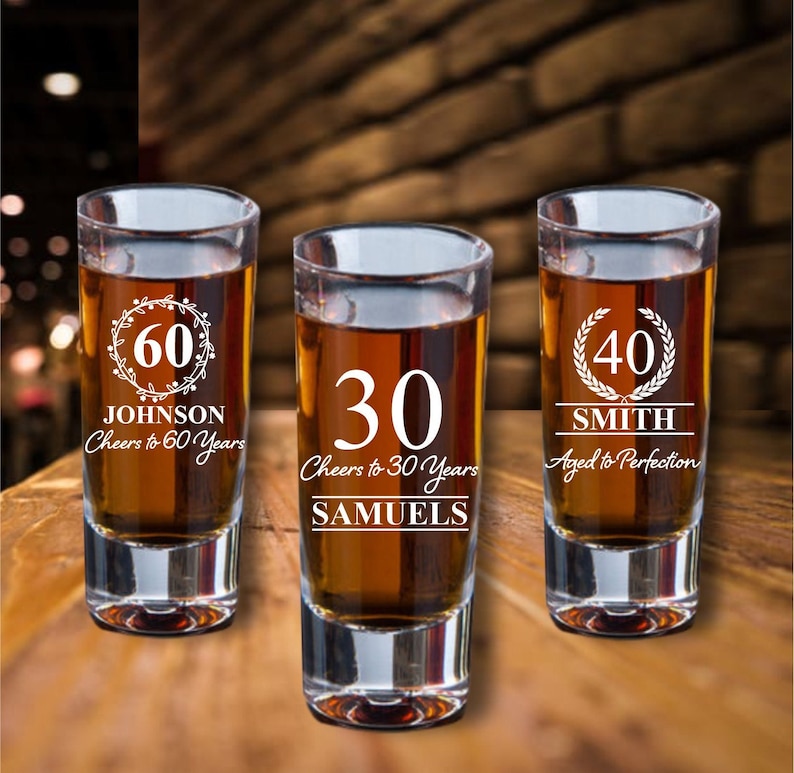 Puede incluir: Tres vasos de chupito de vidrio transparente con grabados personalizados. Los vasos est&aacute;n llenos de un l&iacute;quido &aacute;mbar oscuro. El primer vaso dice "60 JOHNSON Cheers to 60 Years". El segundo vaso dice "30 Cheers to 30 Years SAMUELS". El tercer vaso dice "40 SMITH Aged to Perfection".