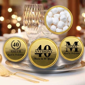 May include: Three gold round favor boxes with black text. The boxes are decorated with a laurel wreath, a number 40, and a large letter M. The text on the boxes reads "40 SMITH Aged to Perfection", "40 ANTHONY Cheers to 40 Years", and "M MIGUEL Aged to Perfection".