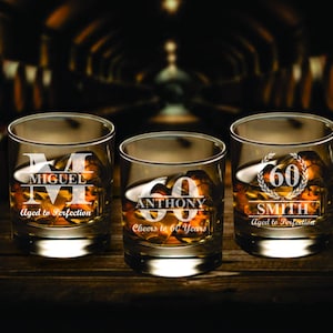 May include: Three personalized whiskey glasses with etched designs. The glasses are filled with amber liquid and have the names "Miguel", "Anthony", and "Smith" etched on them. The glasses are on a wooden surface.