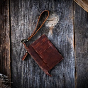 May include: A brown leather wallet with a zipper closure and a strap. The wallet is on a wooden surface.