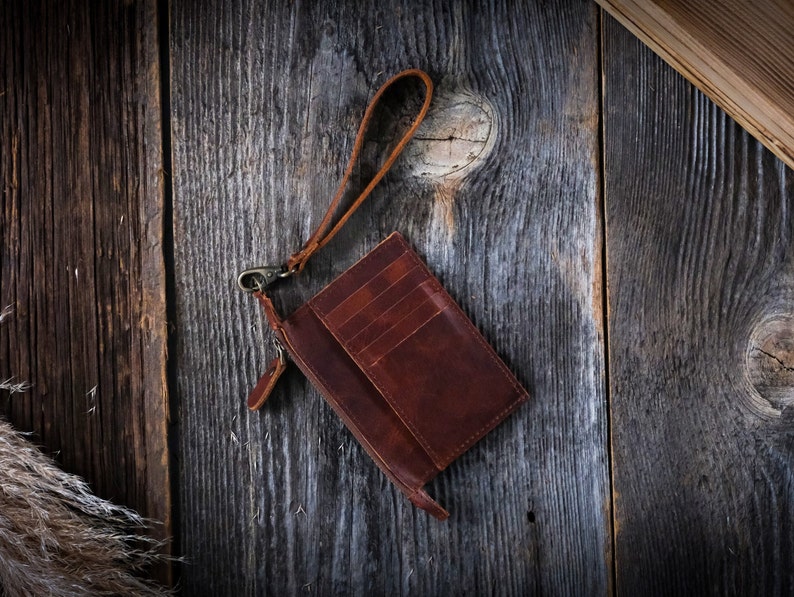 May include: A brown leather wallet with a zipper closure and a strap. The wallet is on a wooden surface.