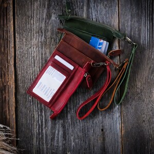 May include: Three leather wallets with different colored straps. The wallets are red, brown, and green. The wallets are on a wooden surface.