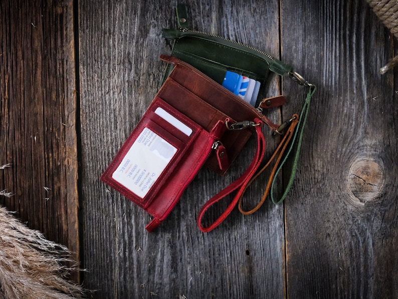 May include: Three leather wallets with different colored straps. The wallets are red, brown, and green. The wallets are on a wooden surface.